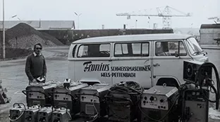 Ein weißer Bus mit der Aufschrift "Fronius Schweißmaschinen Wels-Pettenbach", davor eine Reihe Schweißgeräte